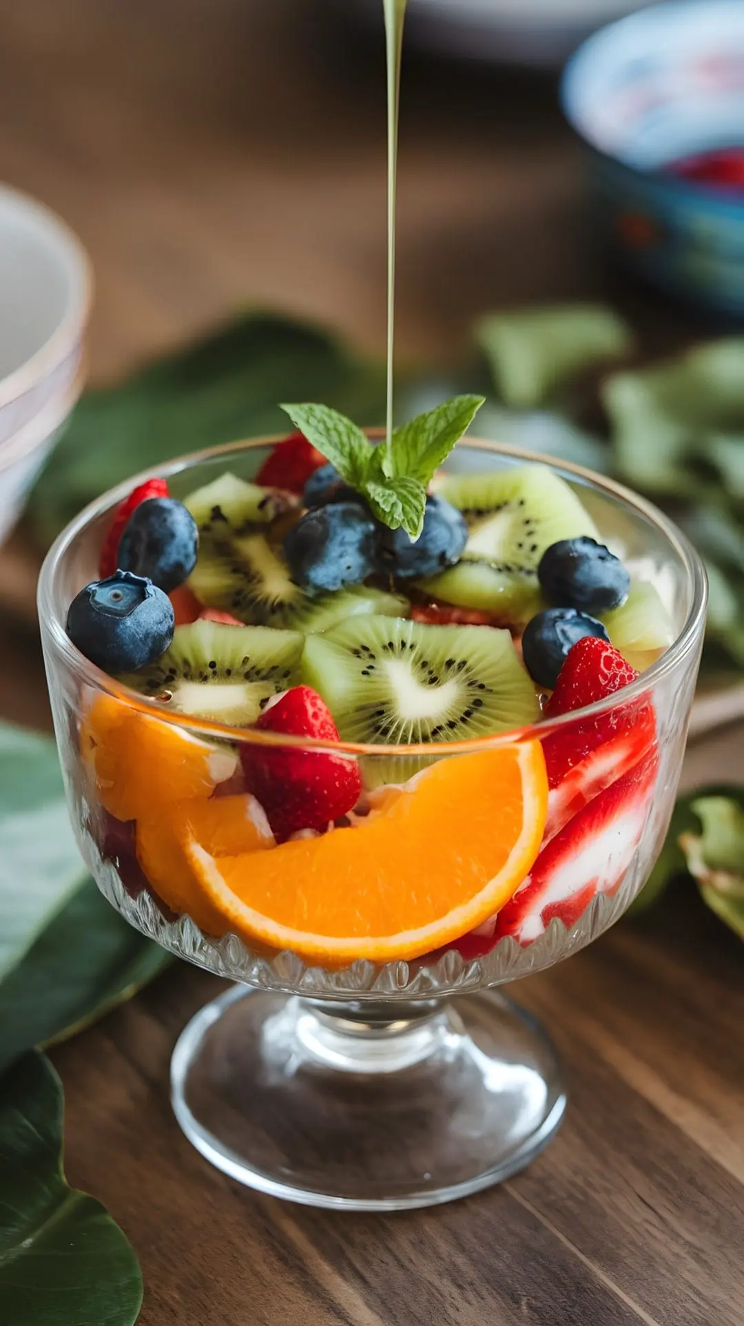 A colorful fruit salad with strawberries, blueberries, kiwi, and orange slices, drizzled with mint dressing, served in a clear glass bowl.